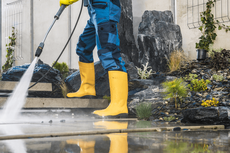a man wearing yellow rubber boots, pressure washing the garden area