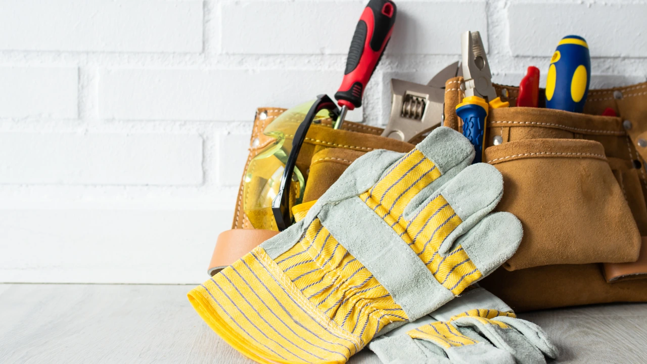 A collection of tools and gloves inside a tool bag is displayed on a wooden floor, representing home maintenance essentials.